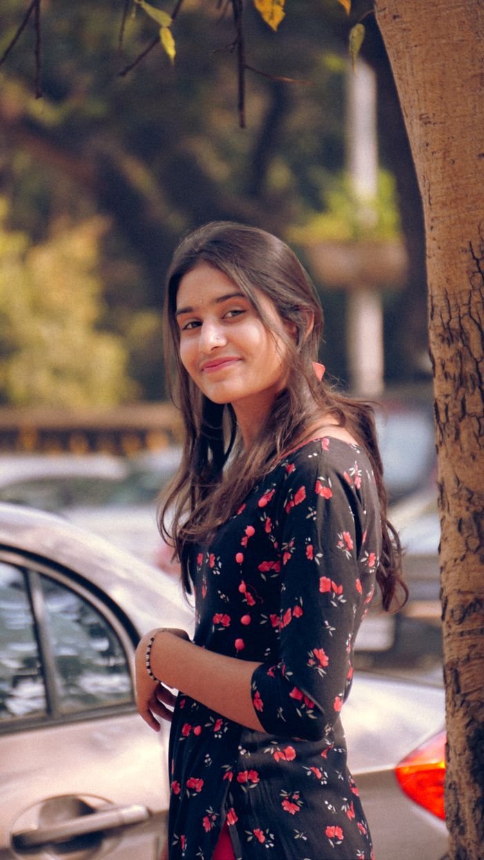 A young woman in a floral dress smiles while standing outdoors beside a car.